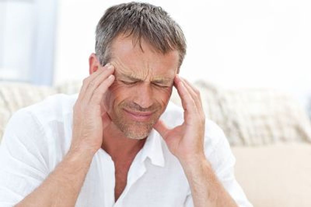 Middle-aged man in discomfort, massaging temples, experiencing a headache, wearing white shirt, seated on couch.