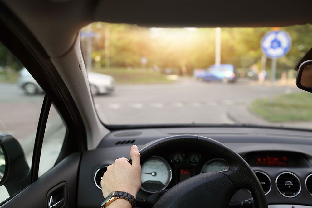 Person driving through a roundabout in daylight, hand on the steering wheel, focus on dashboard and road ahead.