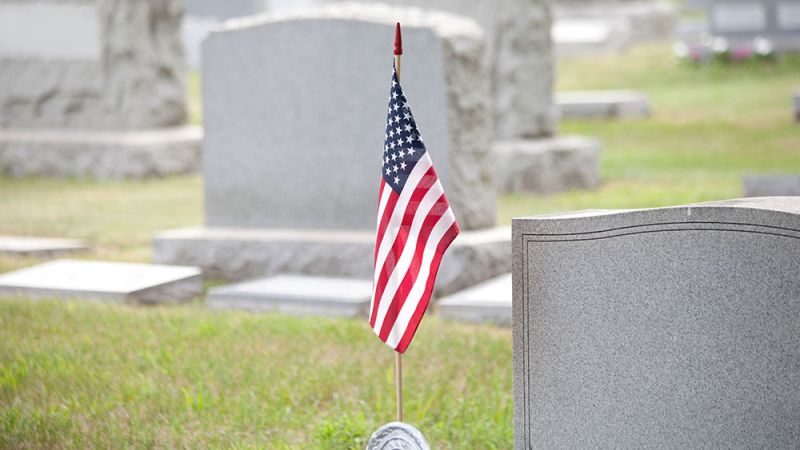 American flag on a grave in a cemetery, surrounded by memorial headstones and green grass.