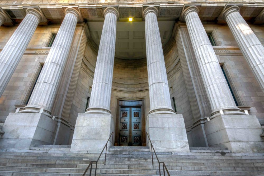 Grand entrance of neoclassical building with towering columns and a massive wooden door, viewed from the staircase.