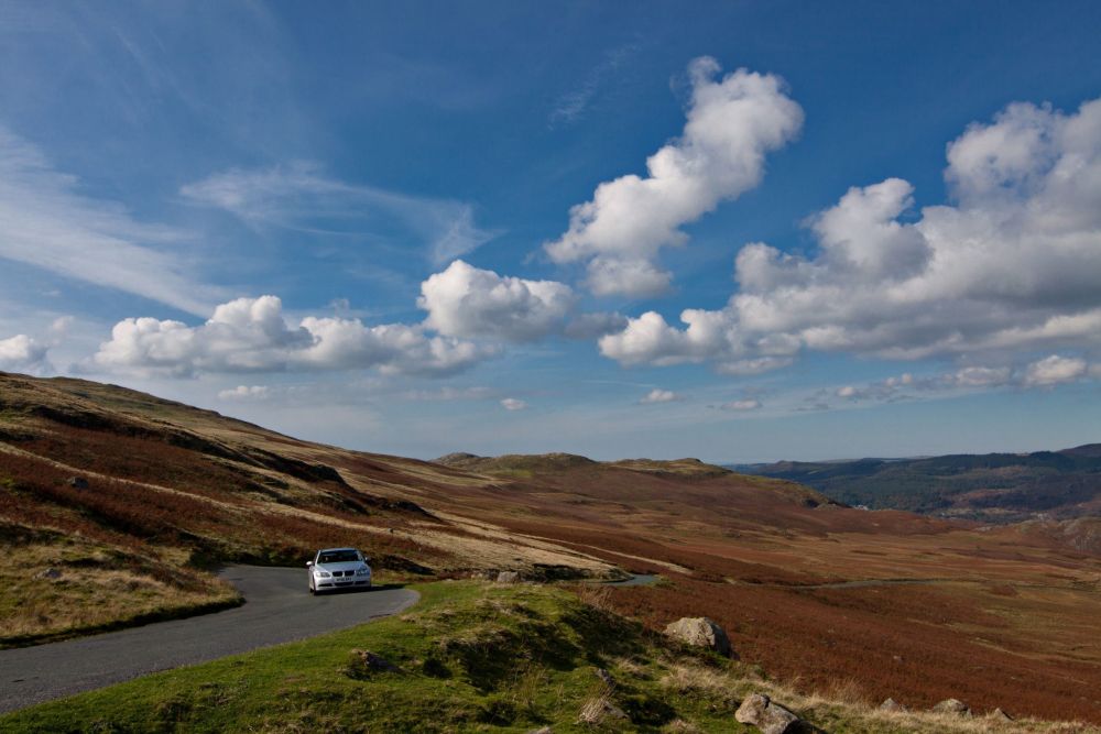 Car on scenic rural road under a sky filled with clouds, surrounded by rolling hills and open landscape.