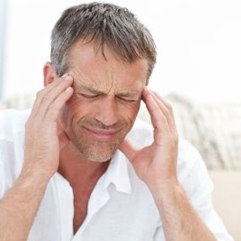 Middle-aged man in discomfort, massaging temples, experiencing a headache, wearing white shirt, seated on couch.