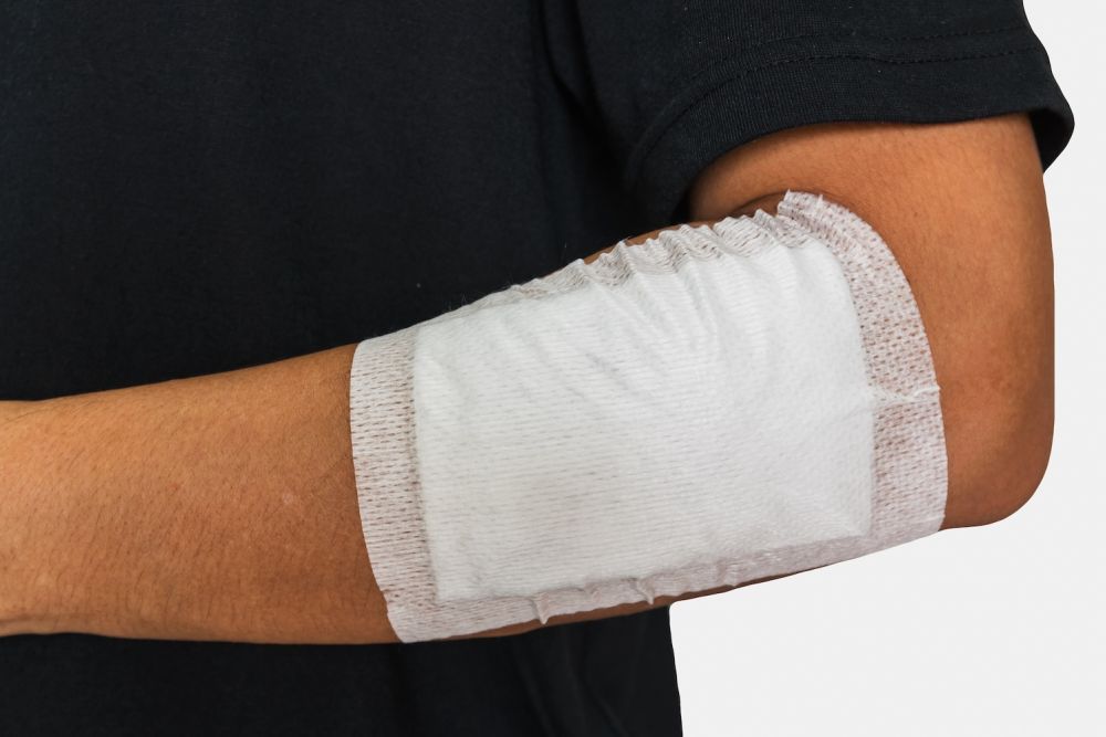 Arm with a bandaged elbow, showing wound care with a white sterile dressing against a black shirt background.