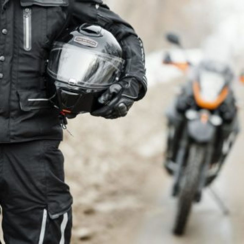 Motorcyclist holding helmet, standing near parked sport bike on a rural road, showcasing safety gear and adventure.