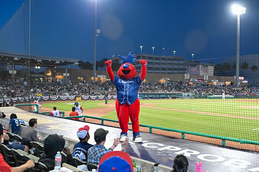 Red baseball mascot entertaining fans at a lively evening game in a brightly lit stadium.