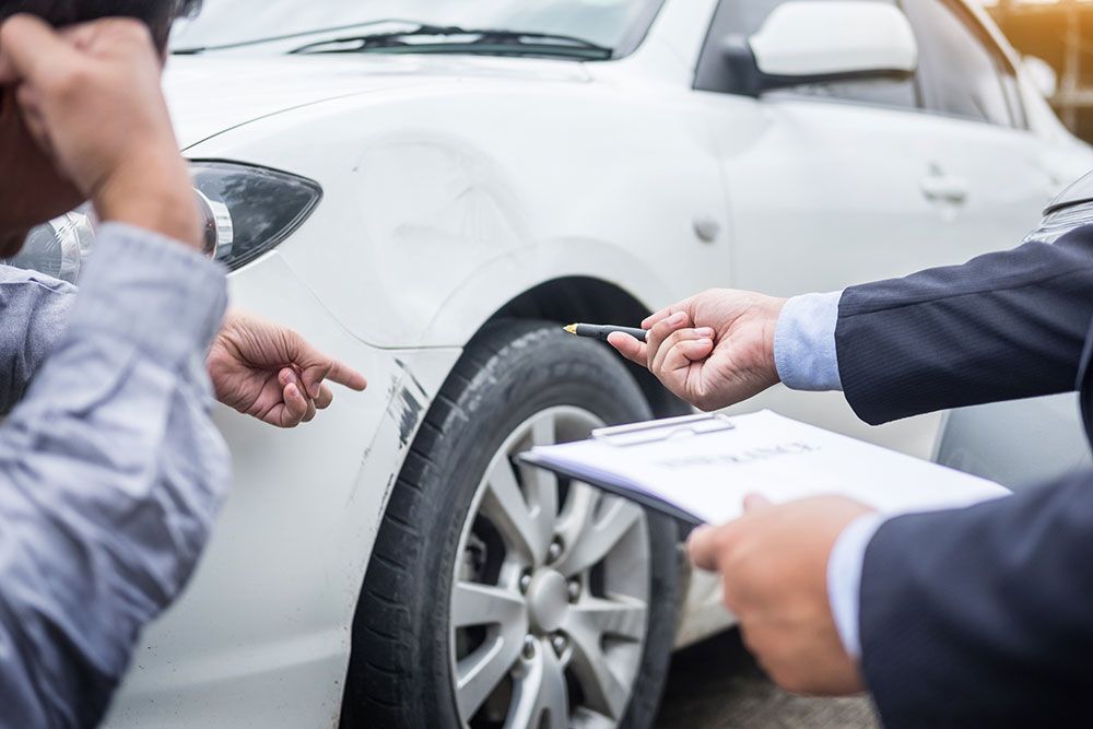 Insurance agent evaluating car damage with a man pointing at a white vehicle's scratched bumper.