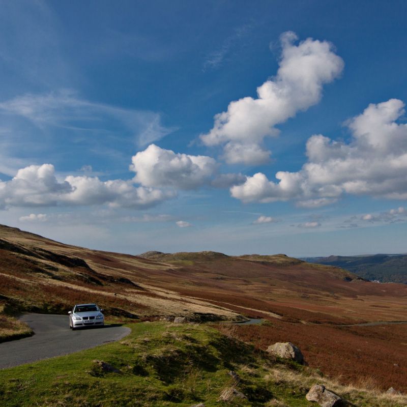 Car on scenic rural road under a sky filled with clouds, surrounded by rolling hills and open landscape.