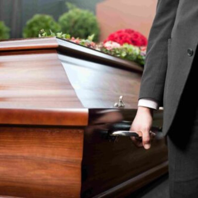 Person in a suit holding a wooden coffin handle adorned with flowers, symbolizing a funeral procession.
