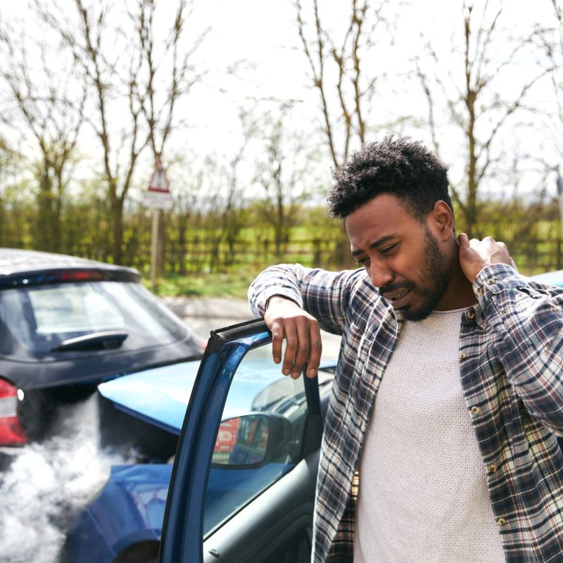 Man in plaid shirt with neck pain after a car accident, standing by a door with smoke in the background.
