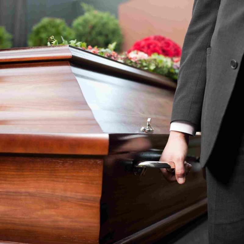 Person holding a wooden casket handle at a funeral, with floral arrangement on top.