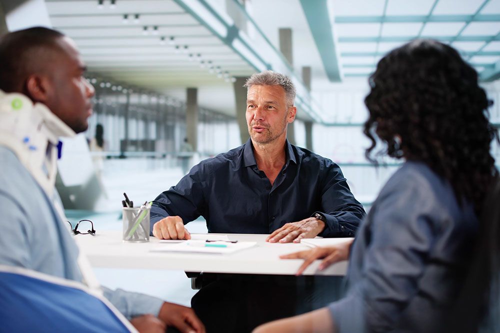 Man in a neck brace consults with a professional at a modern office, discussing an issue with another person present.