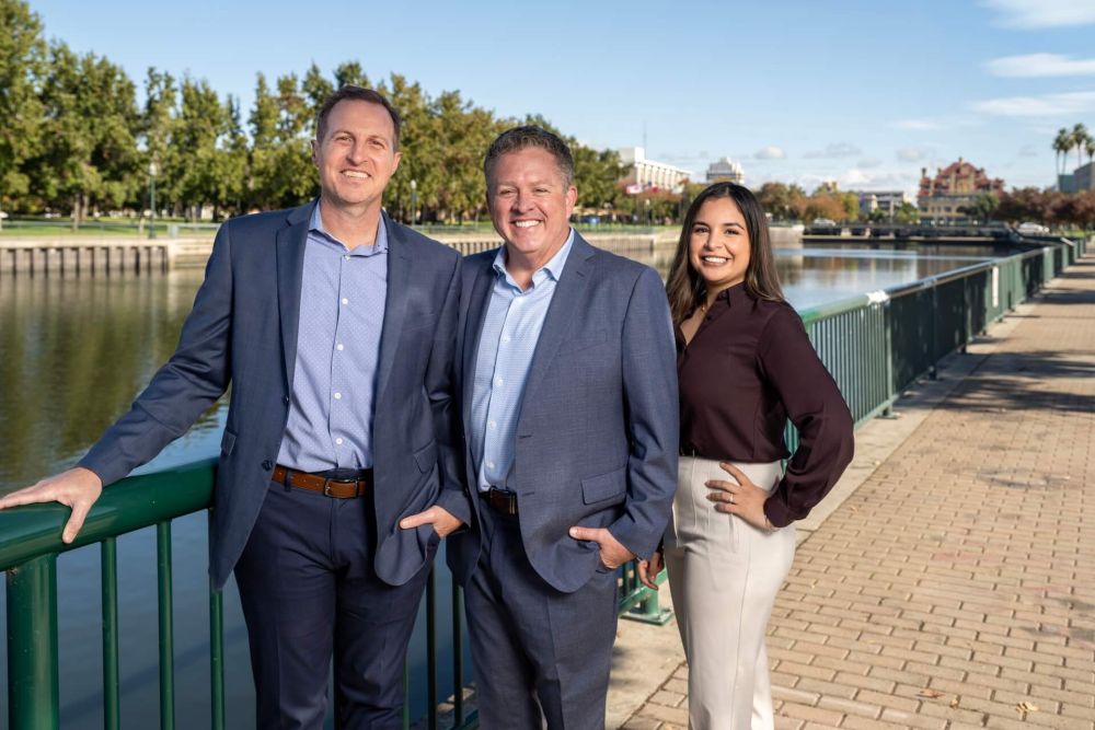 Business professionals smiling by a riverside walkway on a sunny day.