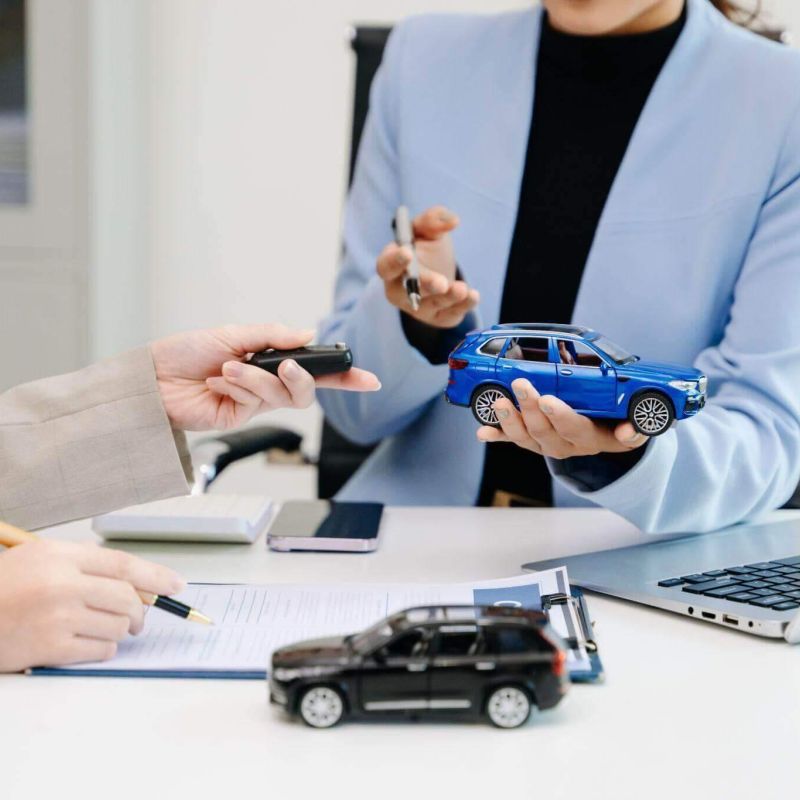 Hands exchange car keys and hold a blue toy car over a desk with paperwork and a laptop, suggesting a business transaction in an office setting.