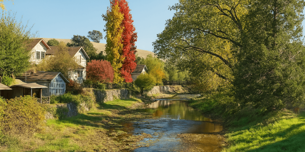 Scenic autumn landscape with colorful trees by a serene creek and rustic houses under a clear blue sky.