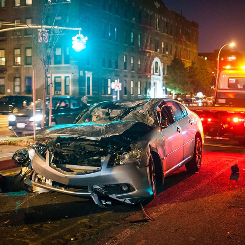 Wrecked silver car on city street at night being towed, traffic and buildings in the background.