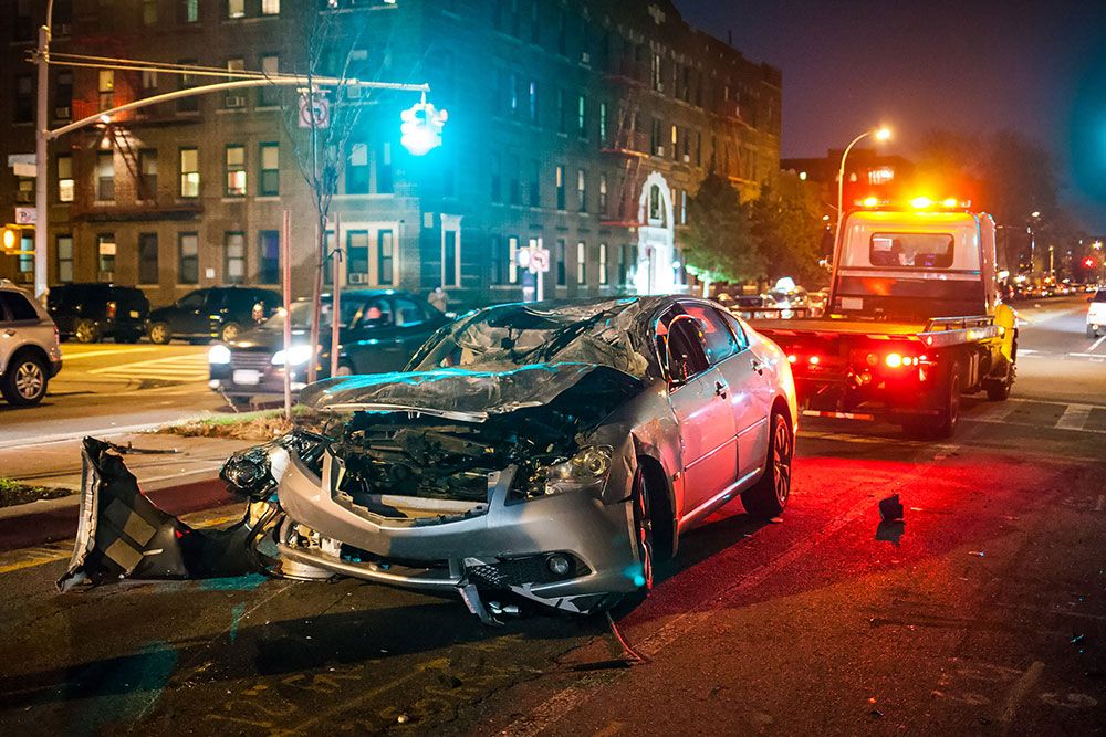 Wrecked silver car on city street at night being towed, traffic and buildings in the background.