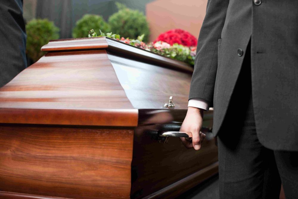 Person holding a wooden casket handle at a funeral, with floral arrangement on top.