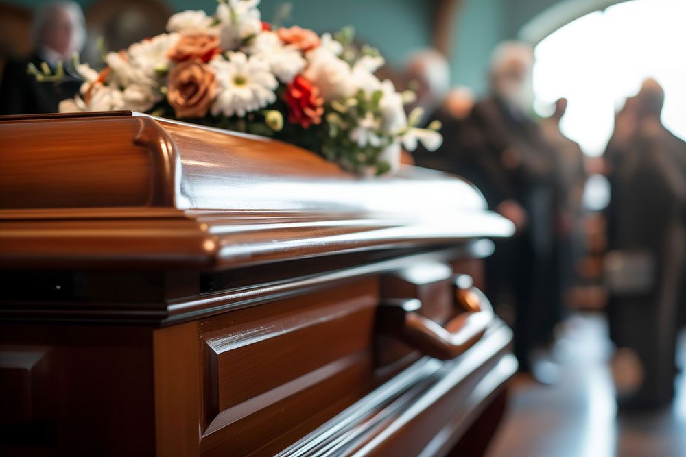 Wooden casket with floral arrangement in a funeral setting, surrounded by blurred attendees.