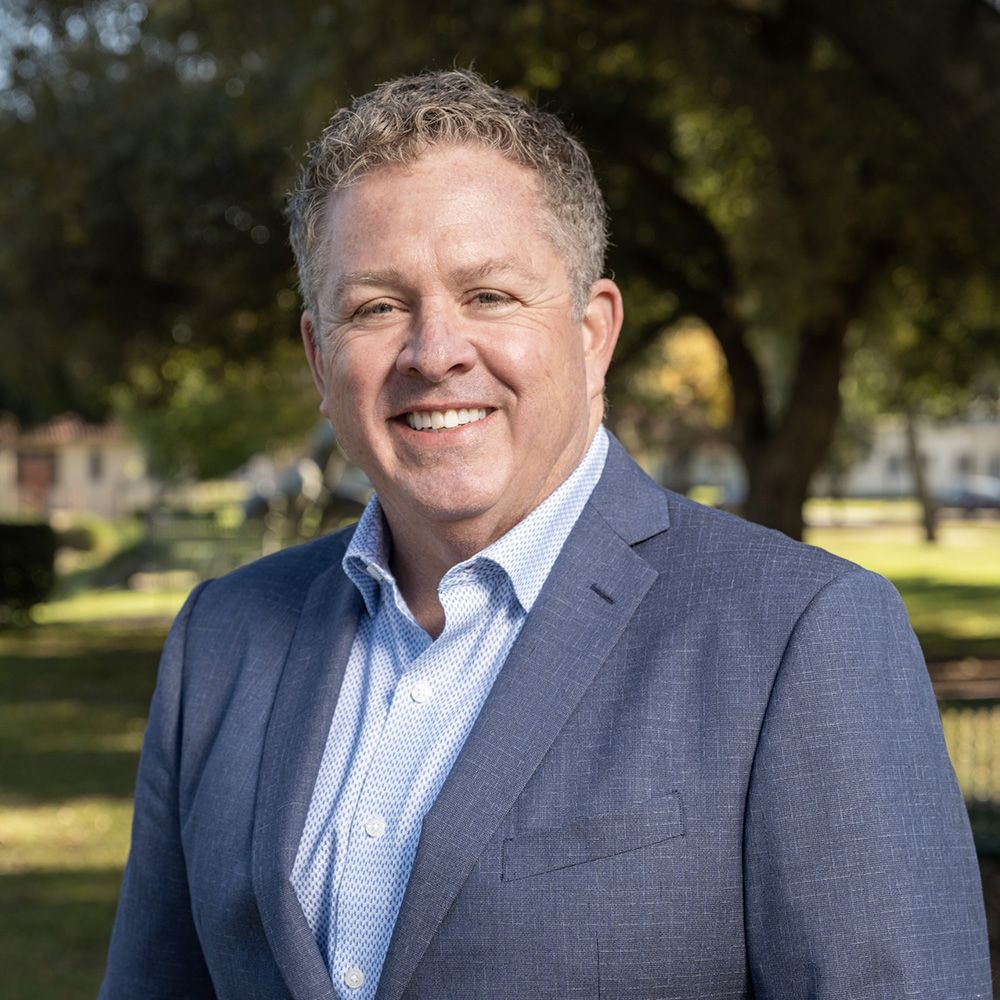 Smiling man in a blue suit standing outdoors with trees in the background.