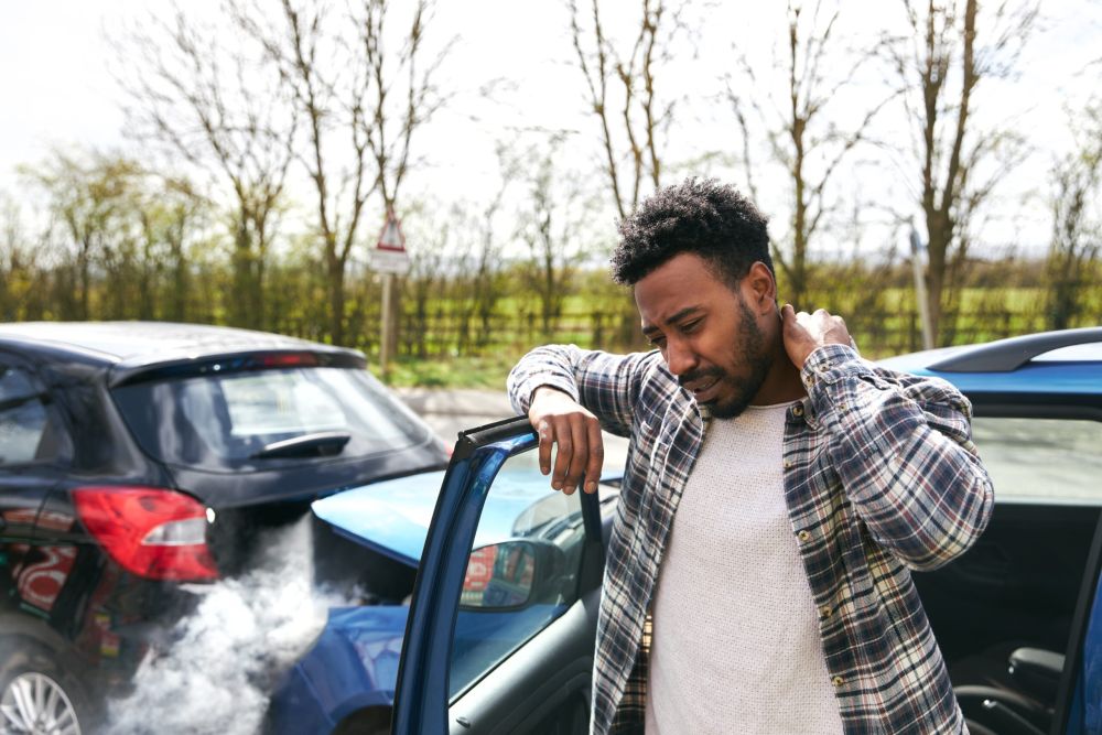 Man in plaid shirt with neck pain after a car accident, standing by a door with smoke in the background.