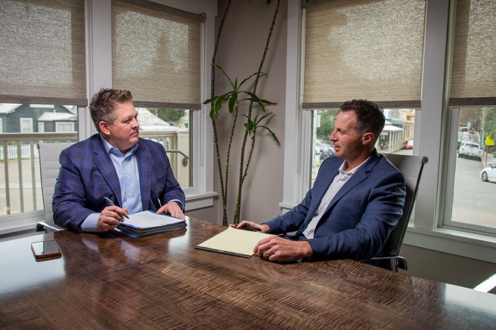 Attorneys Jude Redkey and Robert Gordon have a meeting in a well-lit office, discussing documents on a wooden table.