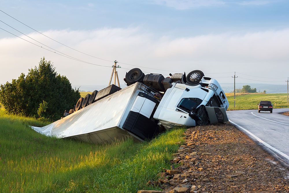 Overturned truck in roadside accident near a rural highway, with another vehicle driving away in the distance.
