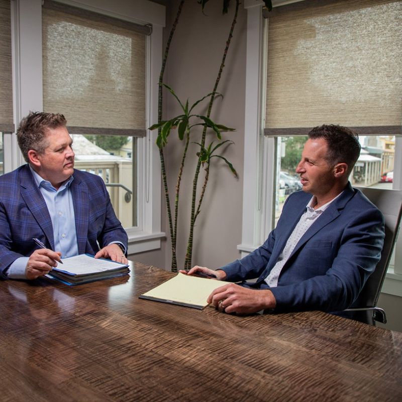 Attorneys Jude Redkey and Robert Gordon have a meeting in a well-lit office, discussing documents on a wooden table.