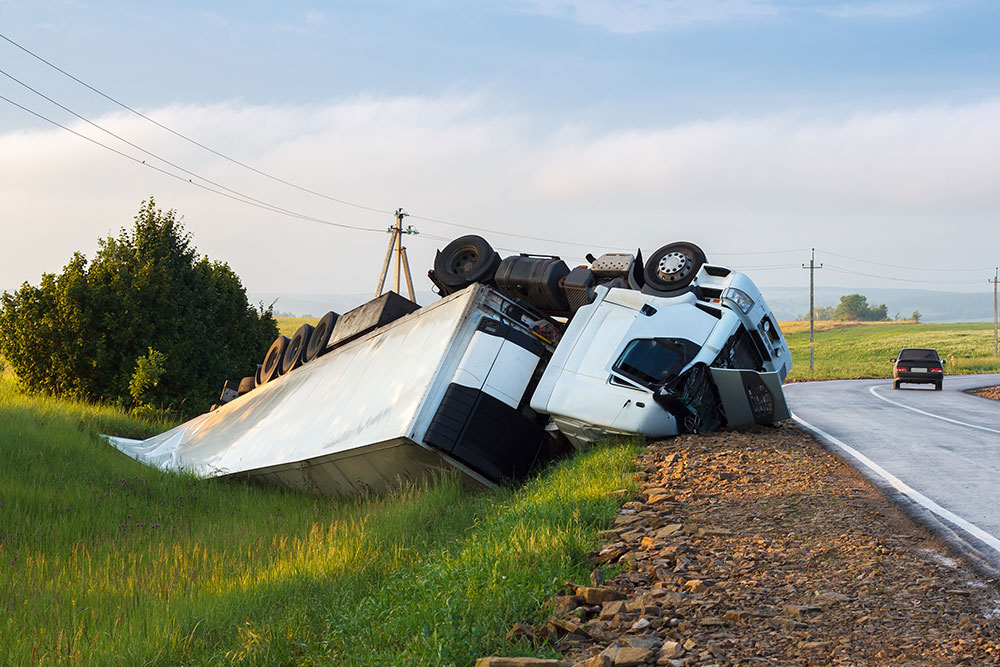 Overturned truck in roadside accident near a rural highway, with another vehicle driving away in the distance.
