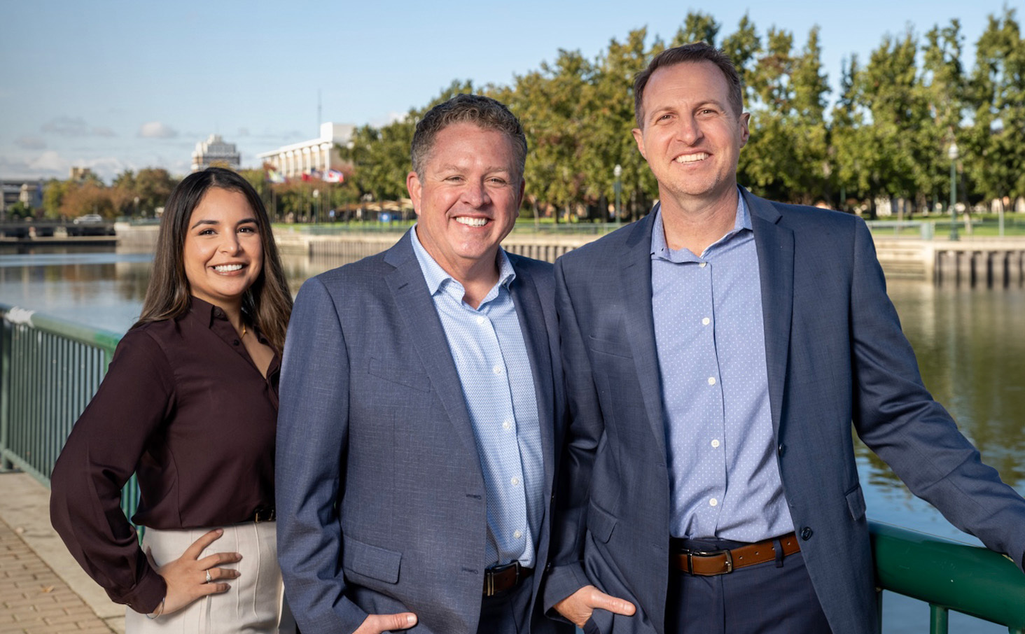 Three professionals smiling in business attire by a waterfront, trees and buildings in the background.