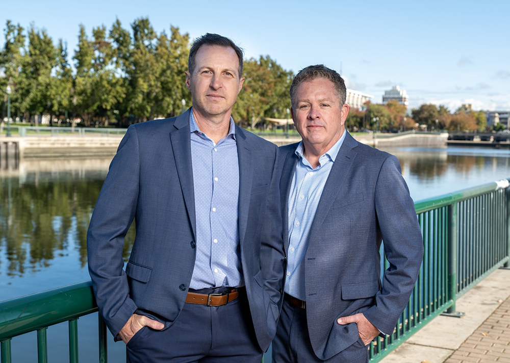 Two businessmen in suits standing by a waterfront railing with trees and buildings in the background.