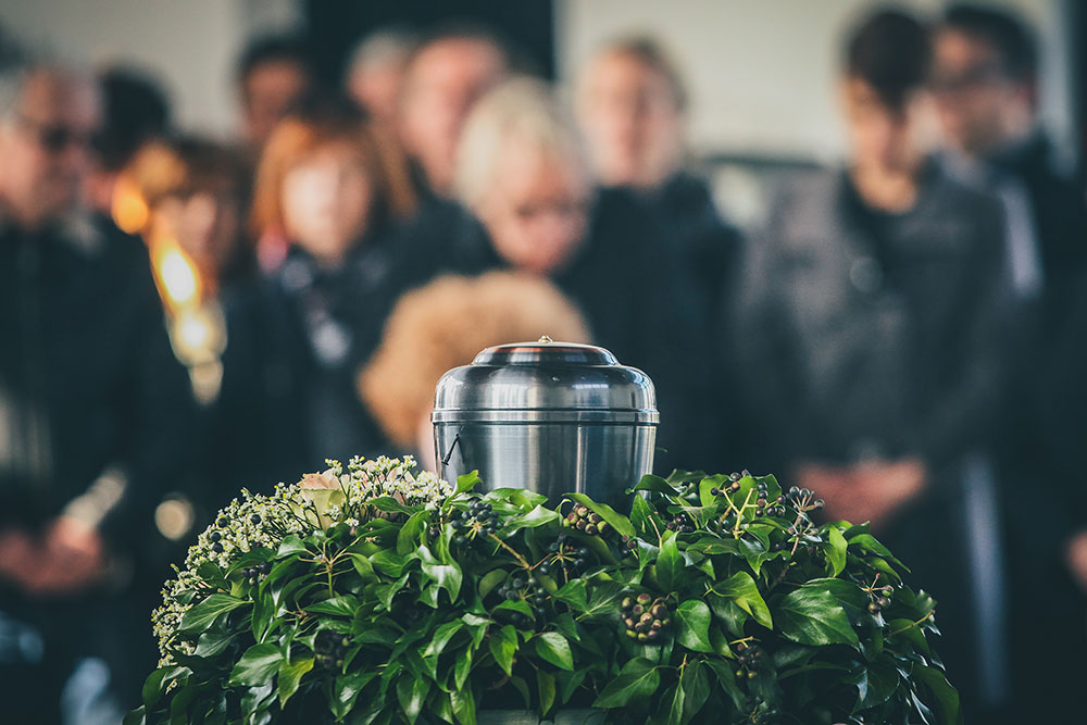 Urn surrounded by flowers at a memorial service with mourners gathered in the background.