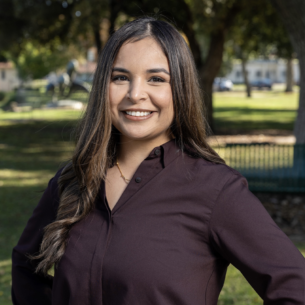 Smiling woman in a dark shirt standing in a sunlit park with trees in the background.