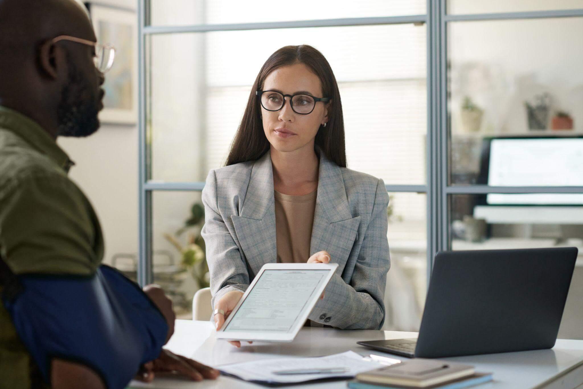 a woman sitting at a desk talking to a man, she is showing him a tablet