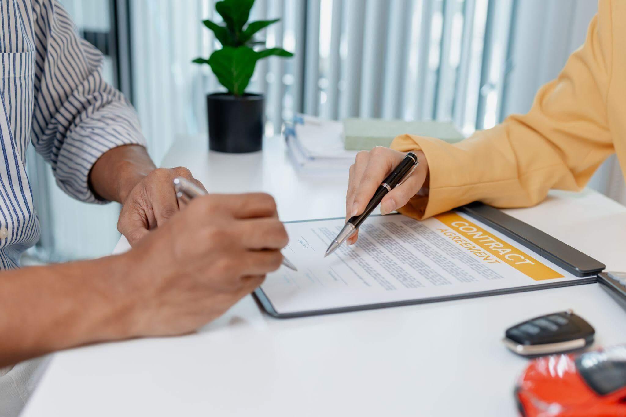 Hands holding pens sign a contract agreement document on a white table. Nearby are a plant, stacked papers, car keys, and a red toy car, with vertical blinds in the background.