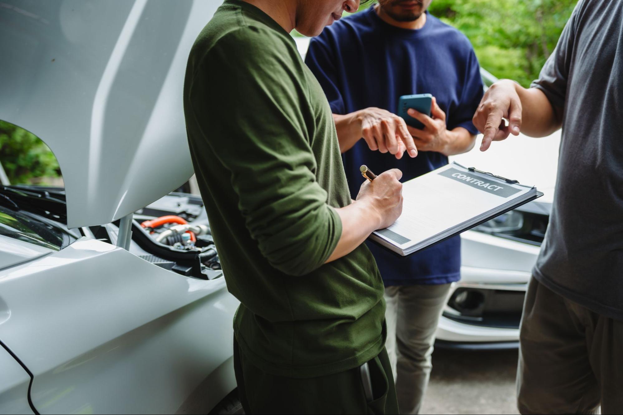 Three people discussing a contract beside an open car hood, one person holding a clipboard and pen.