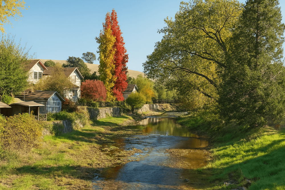Scenic autumn landscape with colorful trees by a serene creek and rustic houses under a clear blue sky.