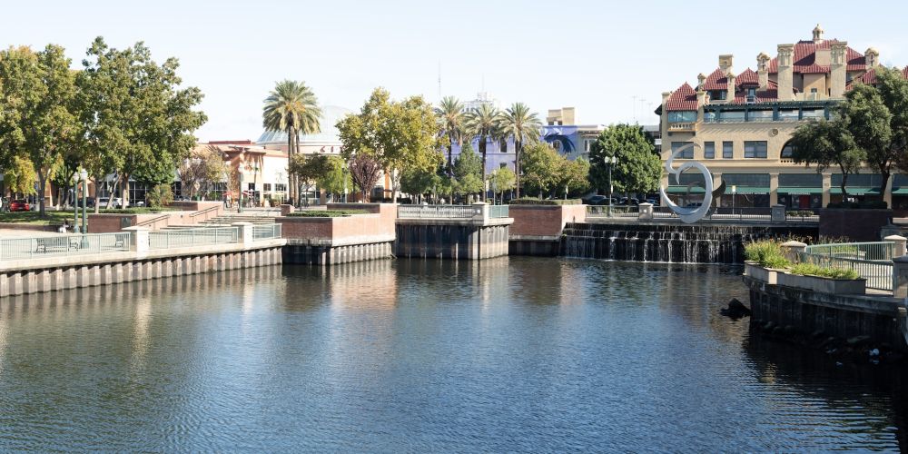 Scenic view of waterfront with trees, buildings, and a circular sculpture under a clear blue sky.