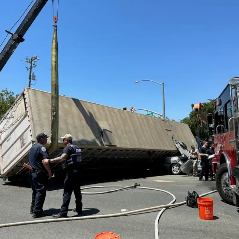 Emergency personnel respond to overturned semi-truck accident with crane and fire truck assistance.