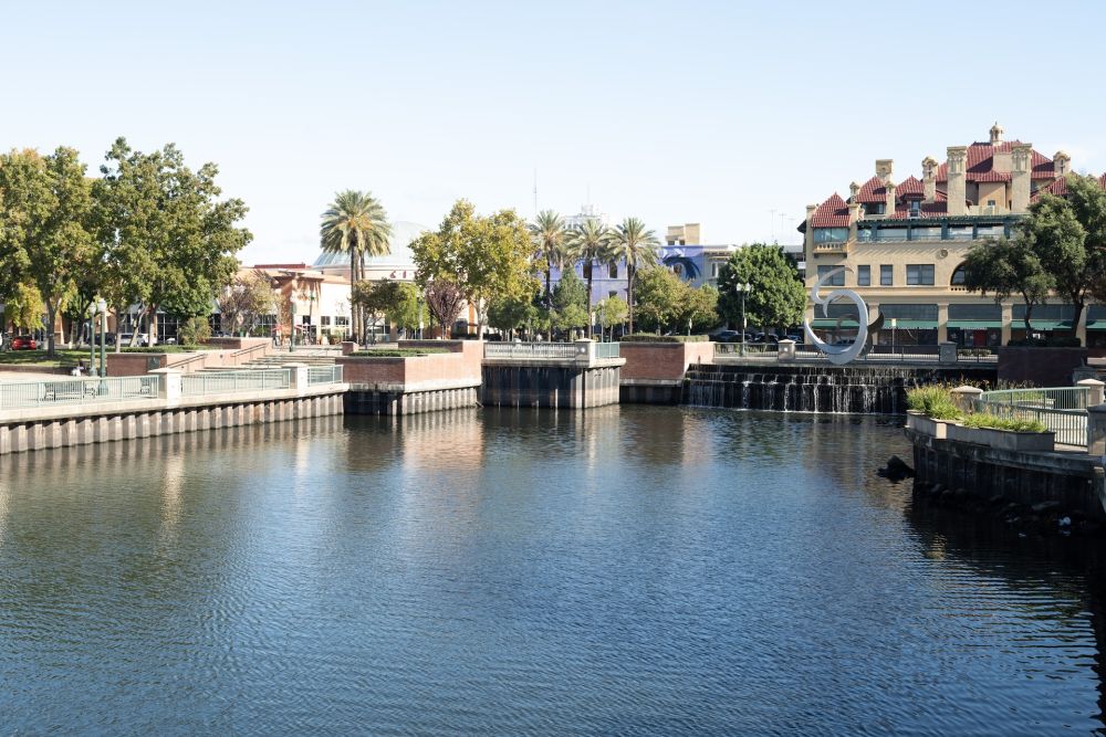 Scenic view of waterfront with trees, buildings, and a circular sculpture under a clear blue sky.