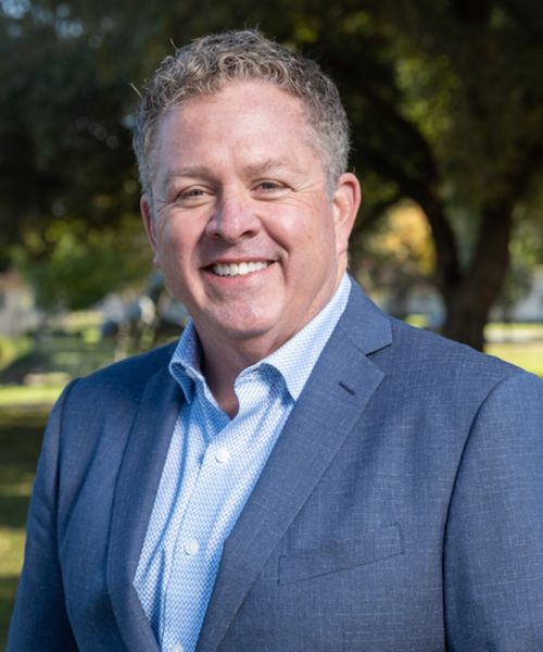 Smiling man in a blue suit outdoors in a park setting, with trees and sunlight in the background.