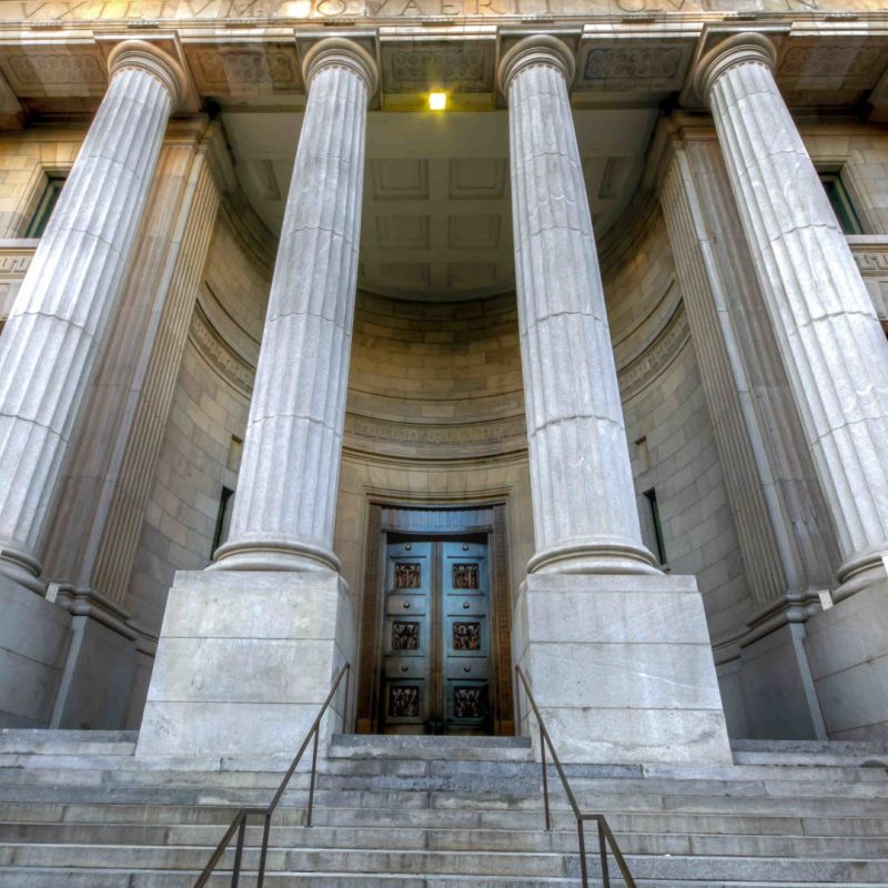 Grand entrance of neoclassical building with towering columns and a massive wooden door, viewed from the staircase.